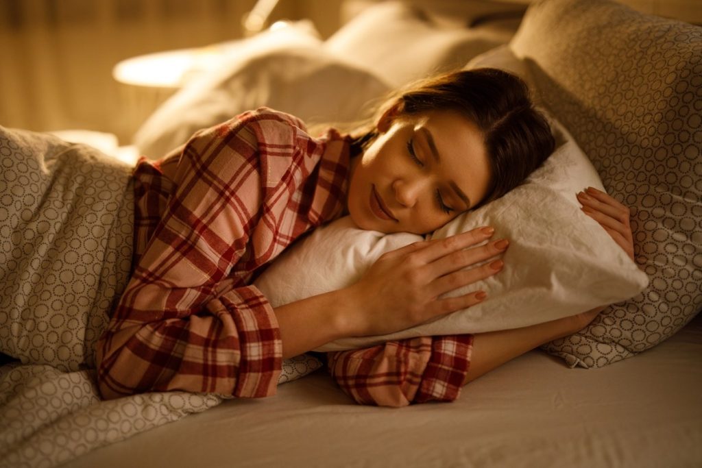 Mulher dormindo na cama abraçada a um travesseiro branco macio. Metrópoles