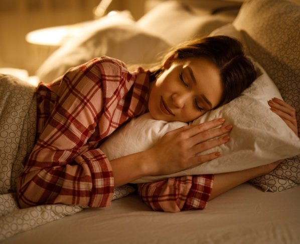 Mulher dormindo na cama abraçada a um travesseiro branco macio. Metrópoles